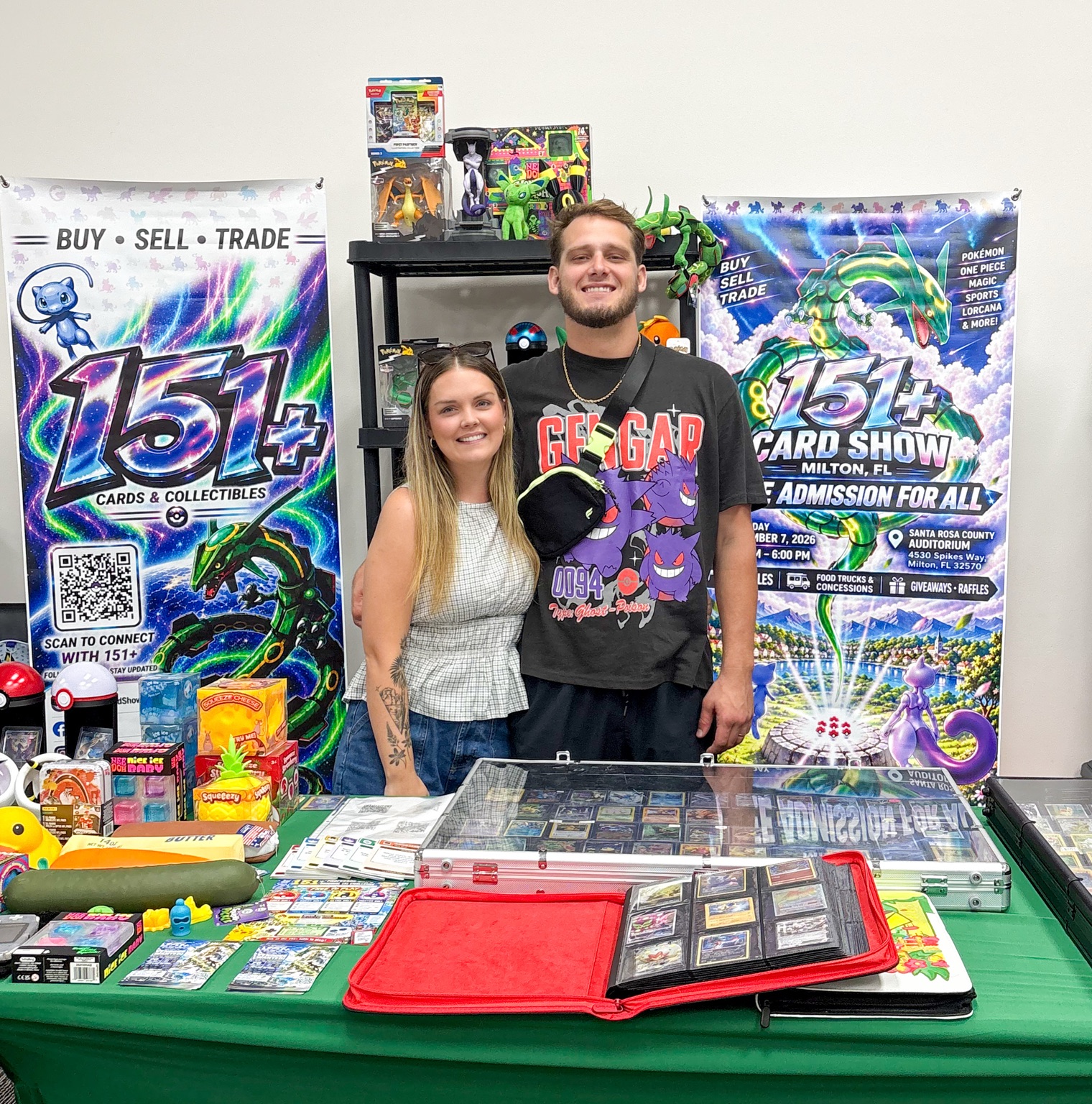 Two collectors smiling behind a display of trading cards and 151 Plus posters